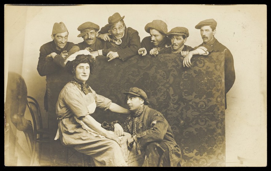 Actors gathered around a man in drag. Photographic postcard by The Fancy Dress Studio, 191-.