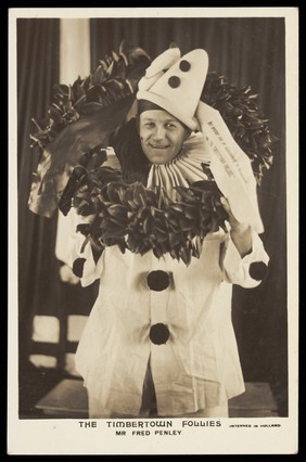 Fred Penley dressed as a clown holding a wreath, performing for "The Timbertown Follies" at a prisoner of war camp in Groningen. Photographic postcard, 191-.