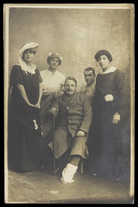 Four soldiers posing, three in drag, with an injured soldier seated in the centre. Photographic postcard, 191-.