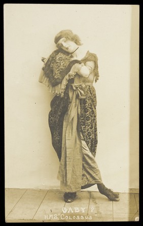 An sailor poses in drag, wearing heavy fabric and a headband. Photographic postcard, ca. 1915-1916.