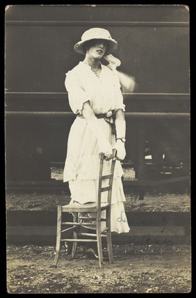 An soldier in a concert party poses in drag, wearing white and holding a chair, standing in front of a train. Photographic postcard, 191-.