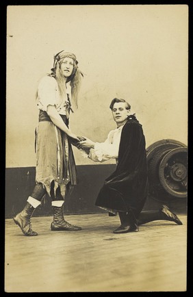 Two amateur actors embrace on the deck of a ship; one is in drag, the other is kneeling on one knee. Photographic postcard, 191-.