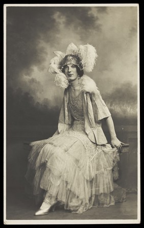 A young man in elaborate drag sits in front of a painted sky backdrop. Photographic postcard, 1934.