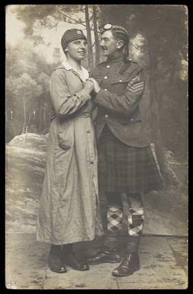 Two soldiers playing in a military concert party embrace: one is in drag and the other is wearing a kilt. Photographic postcard, 191-.