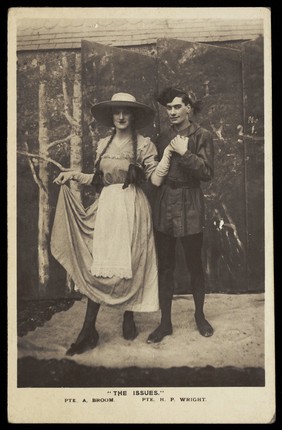 Two members of a military concert party pose on stage: one is in drag with a large hat and pigtails. Photographic postcard, 1915-1916.