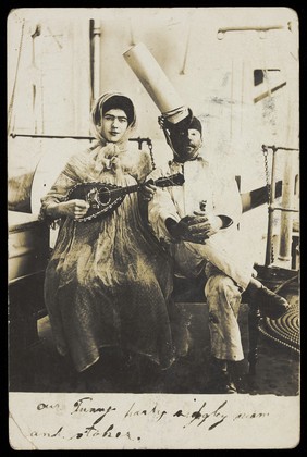 Two sailors (?) on board ship: one in drag is playing a guitar. Photographic postcard, 1907.