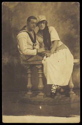 Two sailors, one in drag, embracing while sitting on a stage balcony. Photographic postcard, 191-.