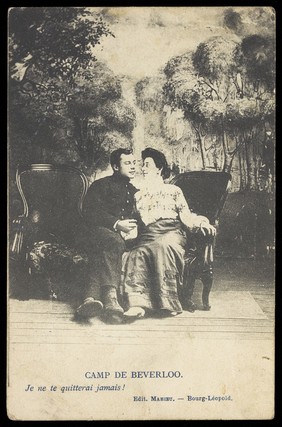 Two soldiers, one in drag, sit close to each other on stage, in front of a detailed backdrop of trees at Beverlo Camp, Belgium. Process print, 190-.