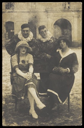 Members of 'The Stars' concert party, two soldiers in drag and two as pierrots, pose around a bench. Photographic postcard, 1919.