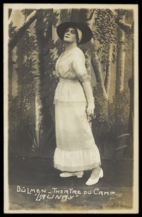 A prisoner of war acting in an internment camp in Dülmen, performing in drag, wearing a white dress and a dark hat. Photographic postcard, 191-.