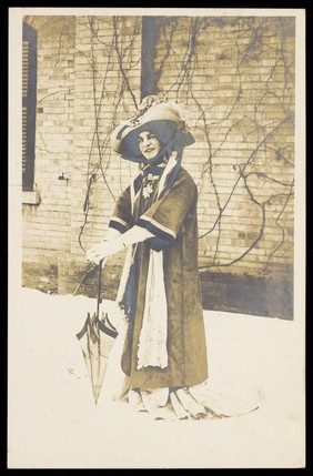 A man in drag, wearing a large bonnet, leans on a parasol whilst standing in front of a brick building. Photographic postcard, 191-.