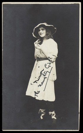 An amateur actor in drag, posing as 'Ninon', wearing a white dress and hat. Photographic postcard, 192-.