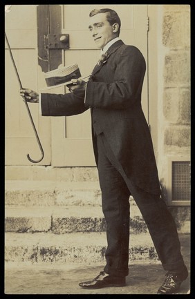 A man wearing a morning suit holds up his cane and boater. Photographic postcard, 191-.