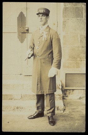 A man dressed wearing a long coat and a hat bearing the word "Cook", stands outside a building. Photographic postcard, 191-.