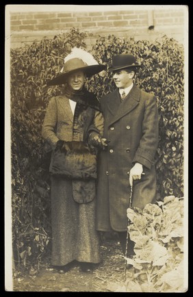 Two men, one in drag, wearing smart attire, pose as a couple within foliage. Photographic postcard, ca. 1900.