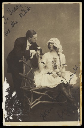 Roy and Gus, two music-hall actors, one in drag, converse across a bench. Photographic postcard by Greenfield Bros, 192-.