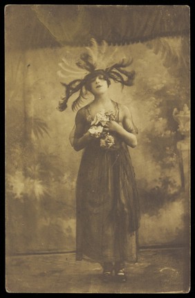 A performer in drag poses on stage wearing a head garment with sprawling feathers. Photographic postcard, 191-.