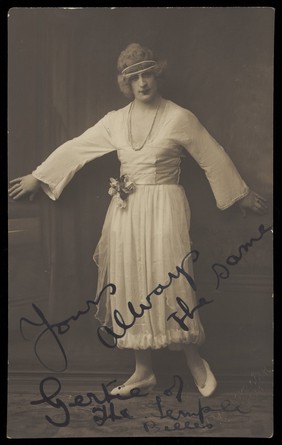 An actor in drag, known as "Gertie", poses mid-movement in a white dress. Photographic postcard, 191-.