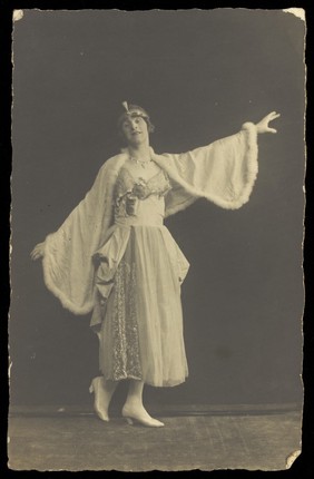 A prisoner of war in drag, wearing a dress and white cloak, poses mid-movement with arms outstretched. Photographic postcard by Albert Melzer, 191-.