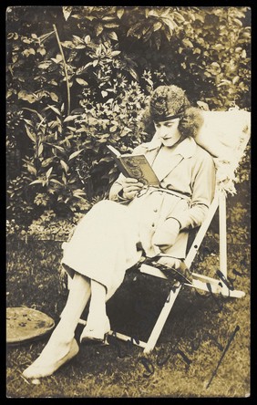 A sailor in drag, wearing a long coat and hat, sits reading in a garden. Photographic postcard, ca. 1920.