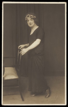 A performer in drag, wearing a black dress and tiara, leaning on a chair. Photographic postcard by W. Aspden, 1924.