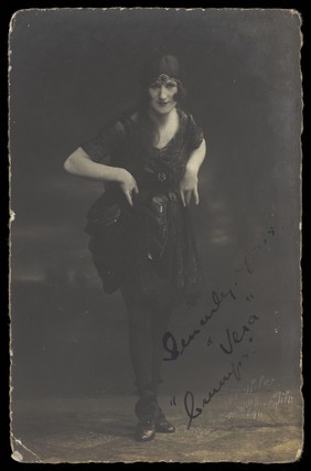 A British prisoner of war in drag, wearing a delicate short dress, leans towards the viewer with his hands on his hips. Photographic postcard by E. Ohle, 191-.