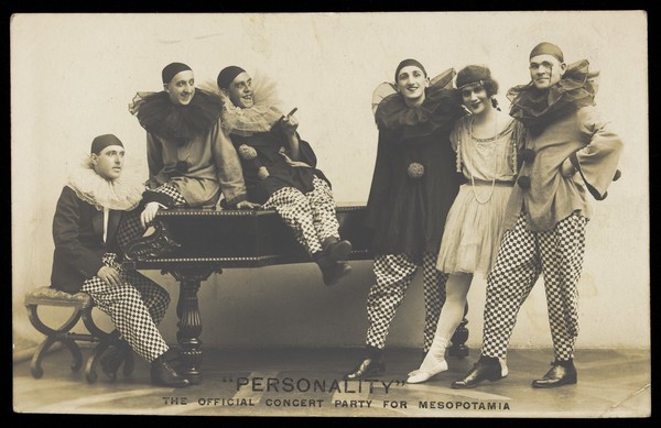 Soldiers, one in drag, performing in the concert party for Mesopotamia, posing around a piano. Photographic postcard by Hana Studios, 191-.