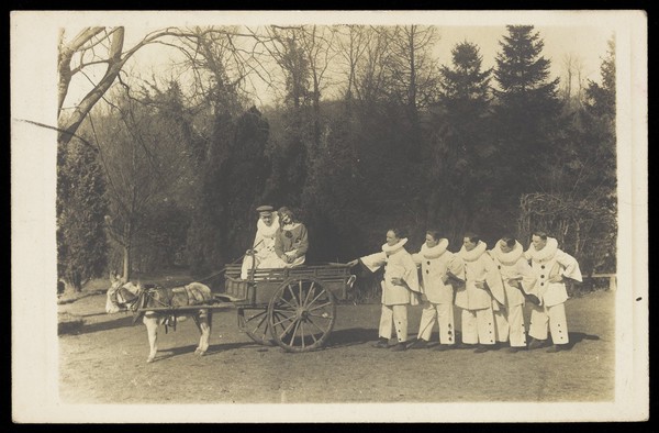 Soldiers, one in drag, with a donkey and cart, posing on the grass in front of trees. Photographic postcard, 191-.