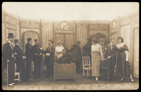 Prisoners of war, some in drag, posing on stage during a crowded scene of "Noblesse oblige"; at Sennelager prisoner of war camp in Germany. Photographic postcard, 191-.