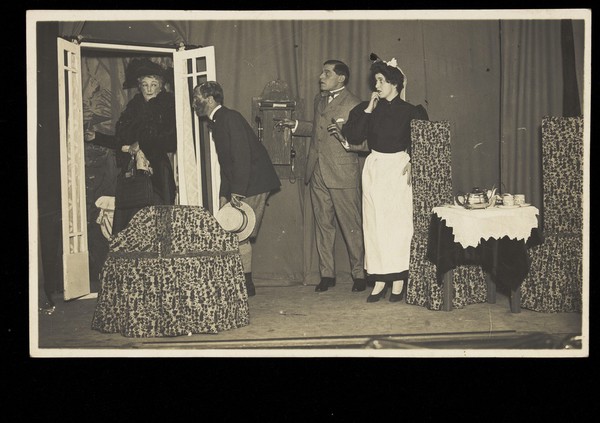 Amateur actors, one in drag, perform a scene on stage. Photographic postcard, 191-.