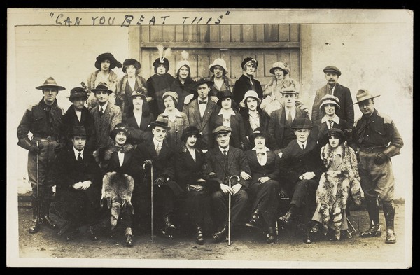 Actors, some in drag, pose for a group portrait, in front of a large wooden doorway. Photographic postcard, 191-.