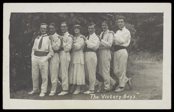 Entertainers, one in drag, pose in a line in front of foliage. Photographic postcard, 191-.