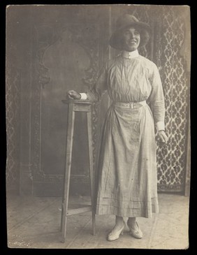 A sailor in drag leans on a high stool, in front of patterned scenery. Photographic postcard, 191-.