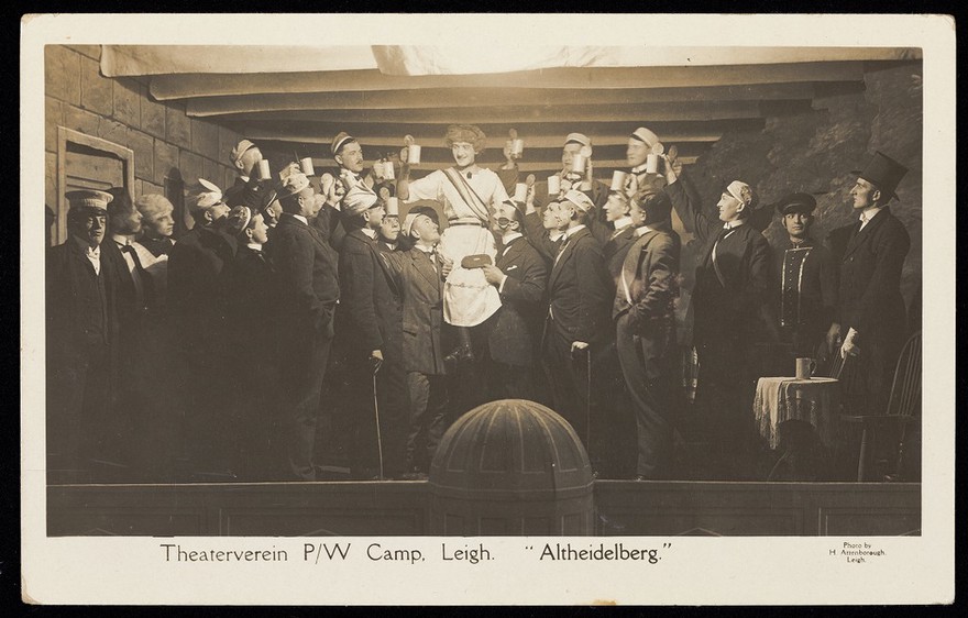 German prisoners of war, one in drag, propose a toast during a performance at Leigh prisoner of war camp, Lancashire. Photographic postcard by H. Attenborough, 191-.