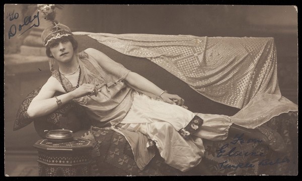 A man in drag poses wearing delicate attire; curled up on a piece of furniture. Photographic postcard, 191-.