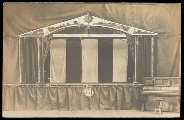A theatre set with a piano beside it and a portrait of a woman mounted at the foot of the stage. Photographic postcard, 191-.