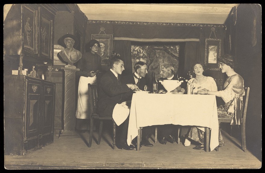Belgian prisoners of war, some in drag, sit at a table within a very detailed stage set at Friedberg camp, Germany. Photographic postcard, 191-.