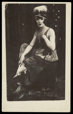 Chris Bennett in drag sits on stage, wearing a feathered head garment. Process print, 1921.