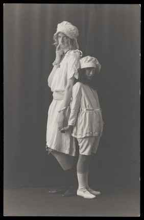 Ralph Mellor in drag and a young actress stand back-to-back on stage. Photographic postcard by L.S. Langfier, 192-.