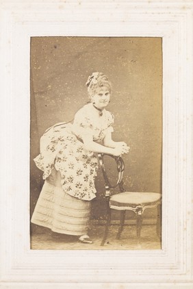 A man in drag resting against a chair. Photograph, 187-.