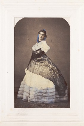 A man in drag poses wearing an elaborate dress. Coloured photograph, 189-.