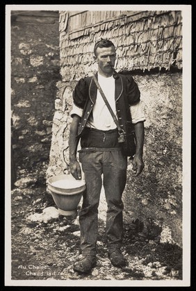 A Swiss farmer standing outside a chalet holding a bucket of fresh milk. Photographic postcard, 19--.