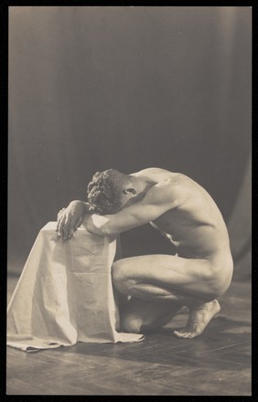 Laurence Woodford, a model, seen from the side, kneeling before a draped pedestal, as if in despair. Photographic postcard, 19--.