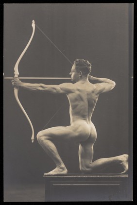 Laurence Woodford, seen in profile, holding a bow and arrow. Photographic postcard, 1928.