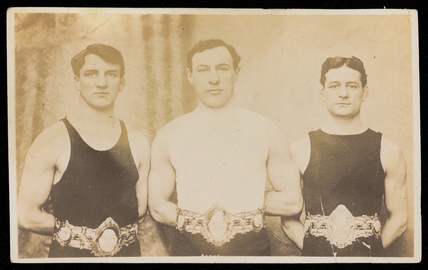 Three male wrestlers posing with champion belts. Photographic postcard, 19--.