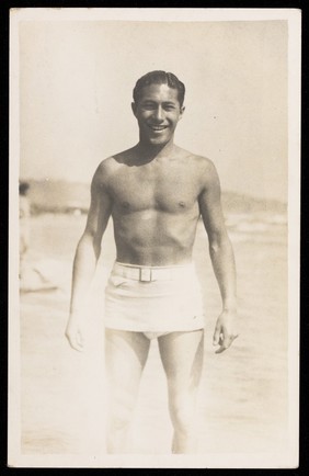 A man stands on a beach wearing a white bathing costume. Photographic postcard, 193-.