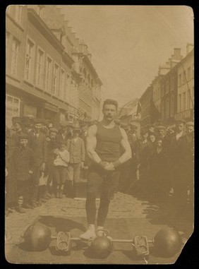 A strongman poses next to his weights surrounded by a crowd. Photograph, 19--.