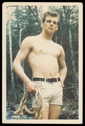 A young man wearing shorts and holding a rope in a wood. Colour photograph, 195-.