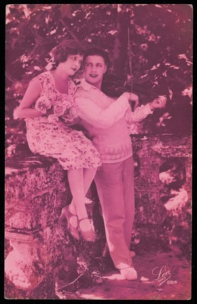 A young woman holding roses sits lovingly next to a young man playing the violin. Coloured photographic postcard, 192-.