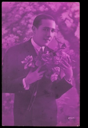 A young man holding roses, engaging the viewer. Colour photographic postcard, 192-.